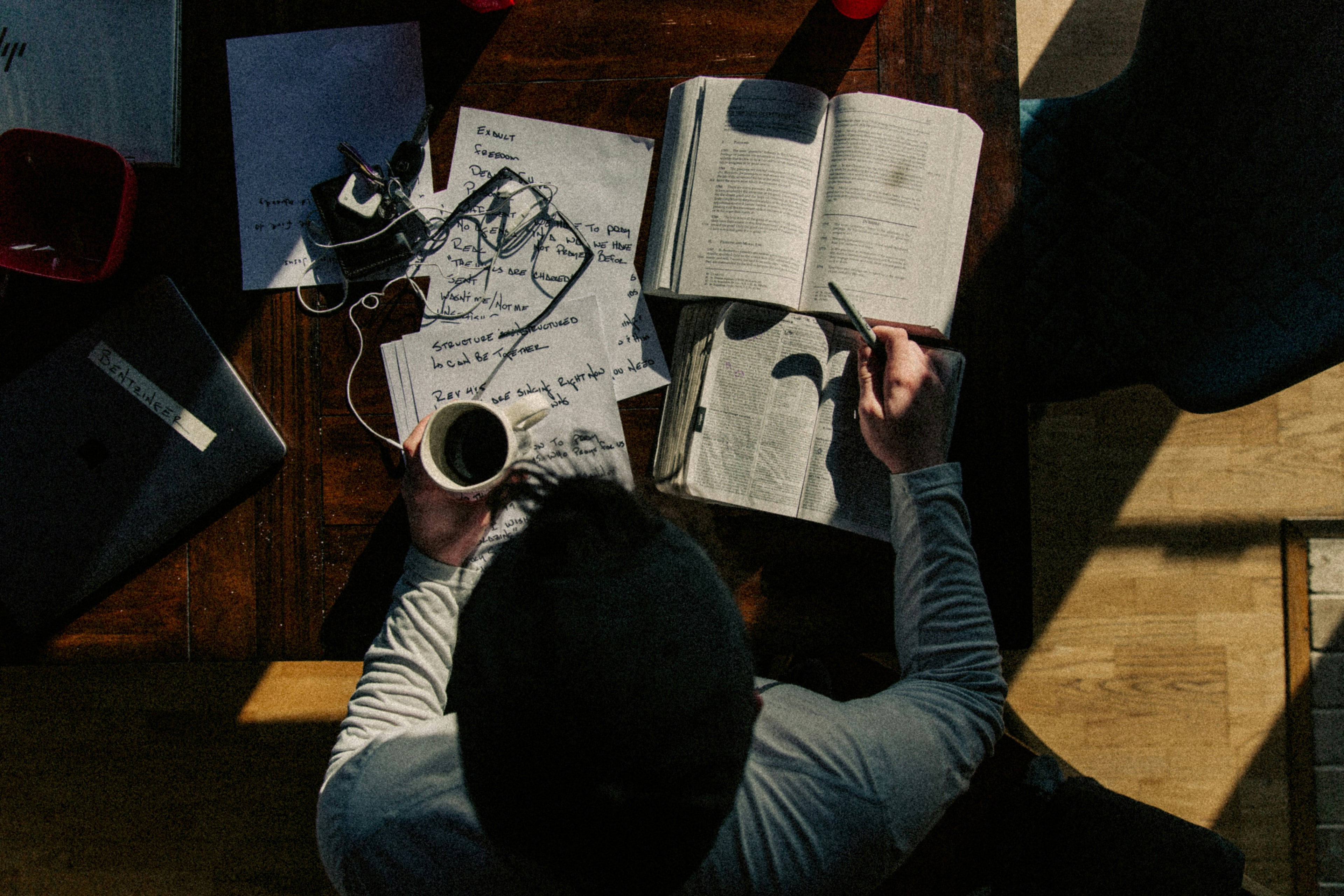 Person sitting at a desk with a notebook, pen, and coffee cup, surrounded by books and papers.